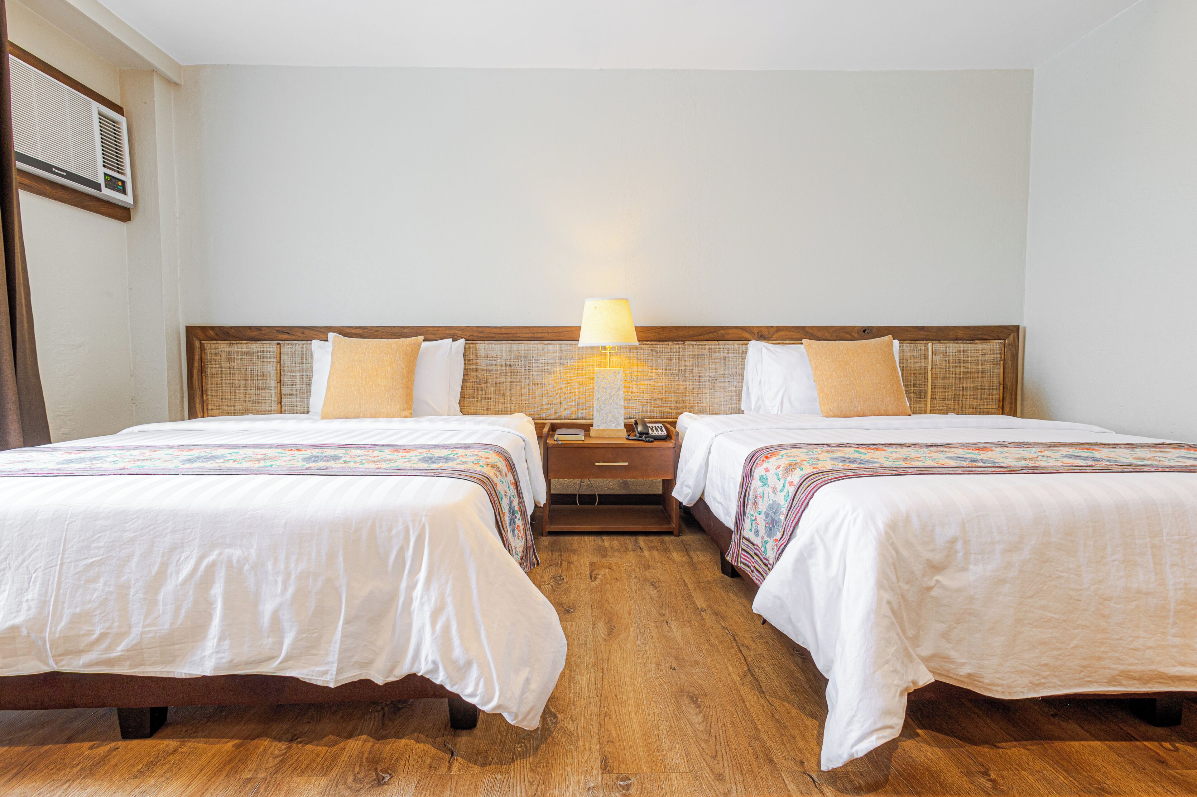 A hotel room with two beds, a bedside table with a lamp, and a wall-mounted air conditioner.