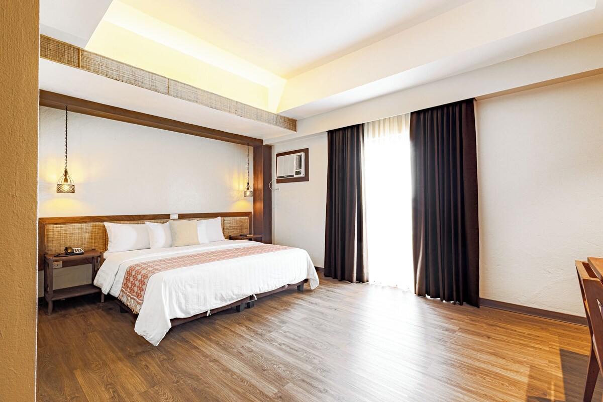 Een hotelkamer met een houten vloer, een bed met wit beddengoed, een nachtkastje, een lamp en een raam met gordijnen.
