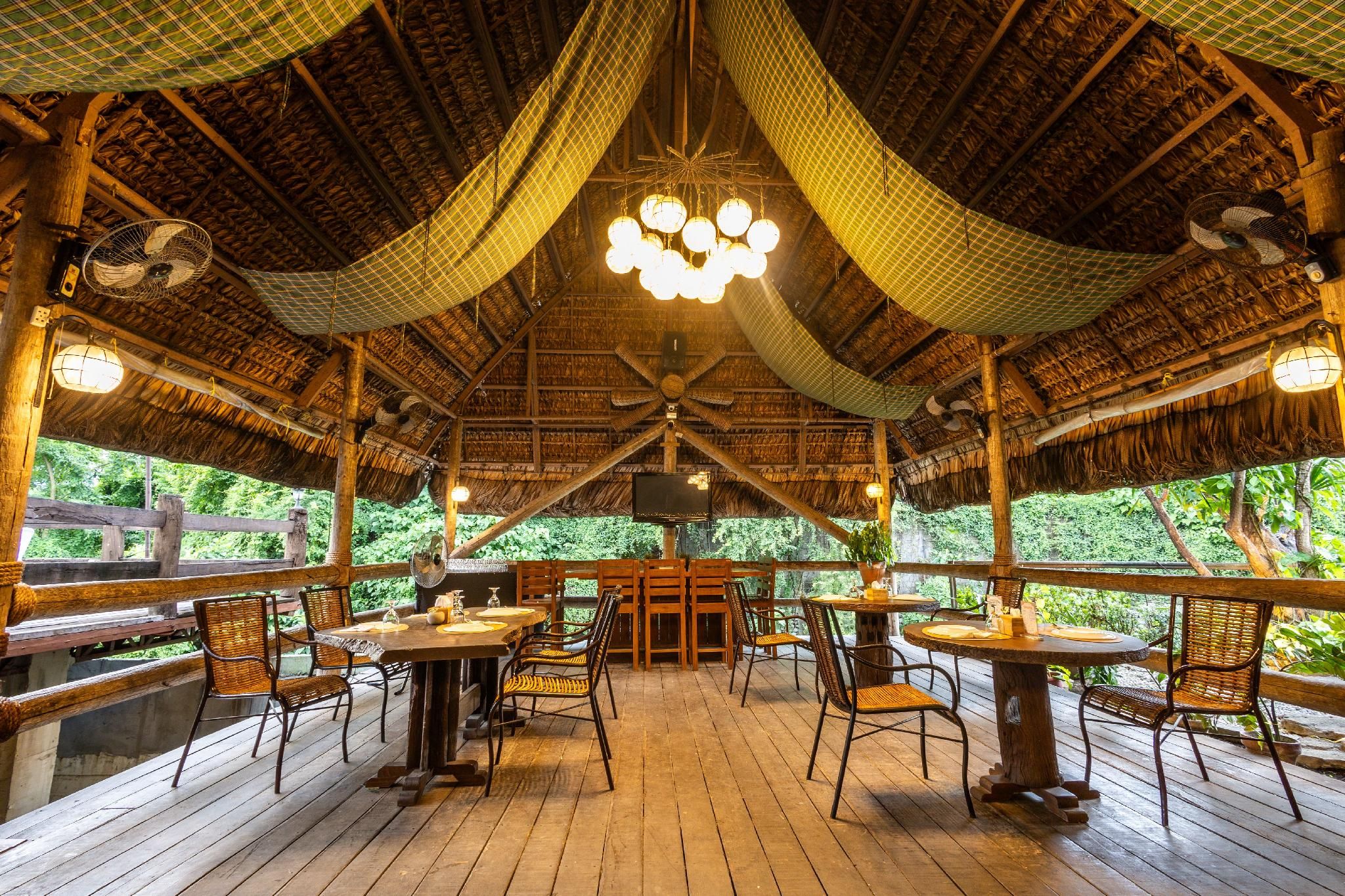 An outdoor dining area with wooden tables and chairs, a thatched roof, and a view of greenery.