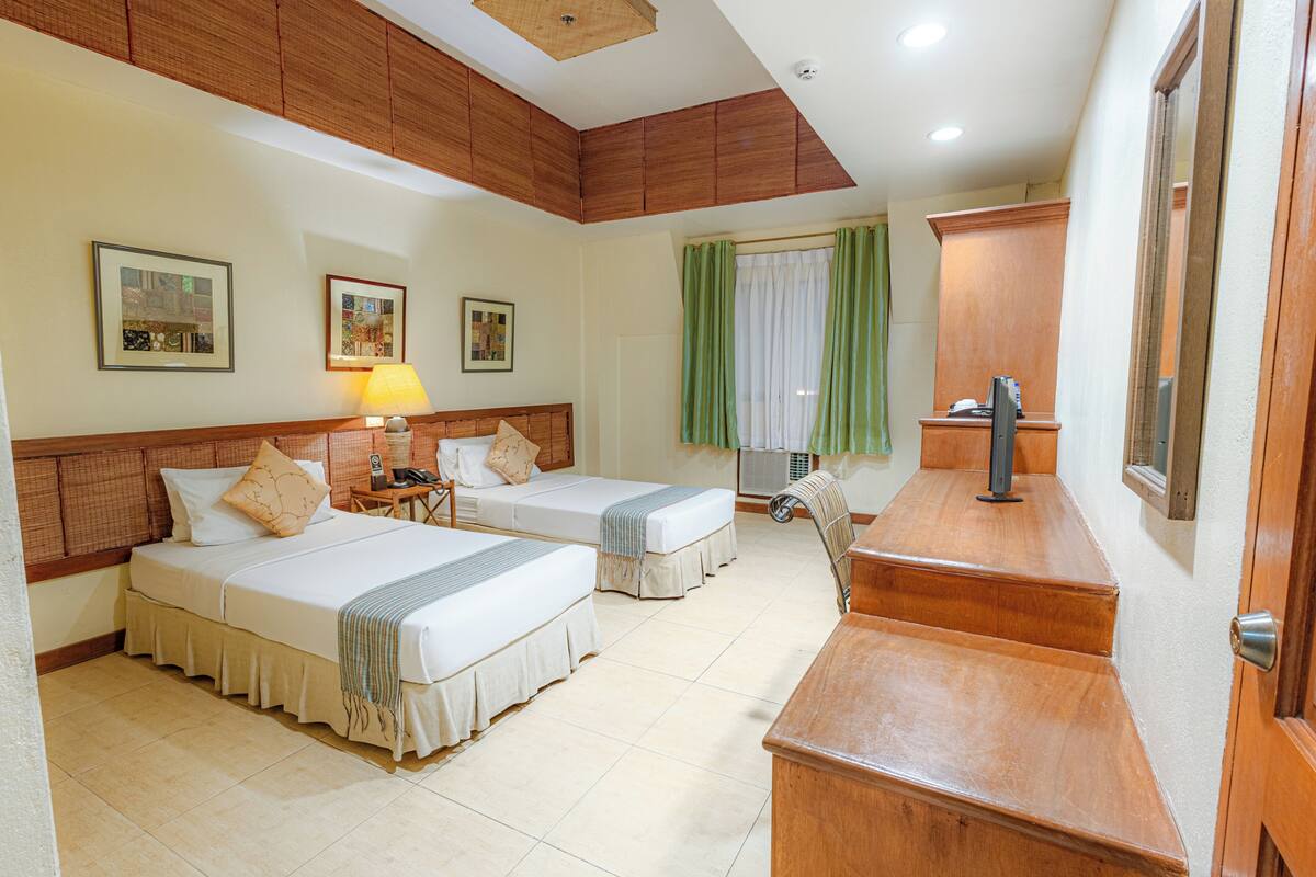 A hotel room with two beds, a desk, a chair, a lamp, and a window with curtains.
