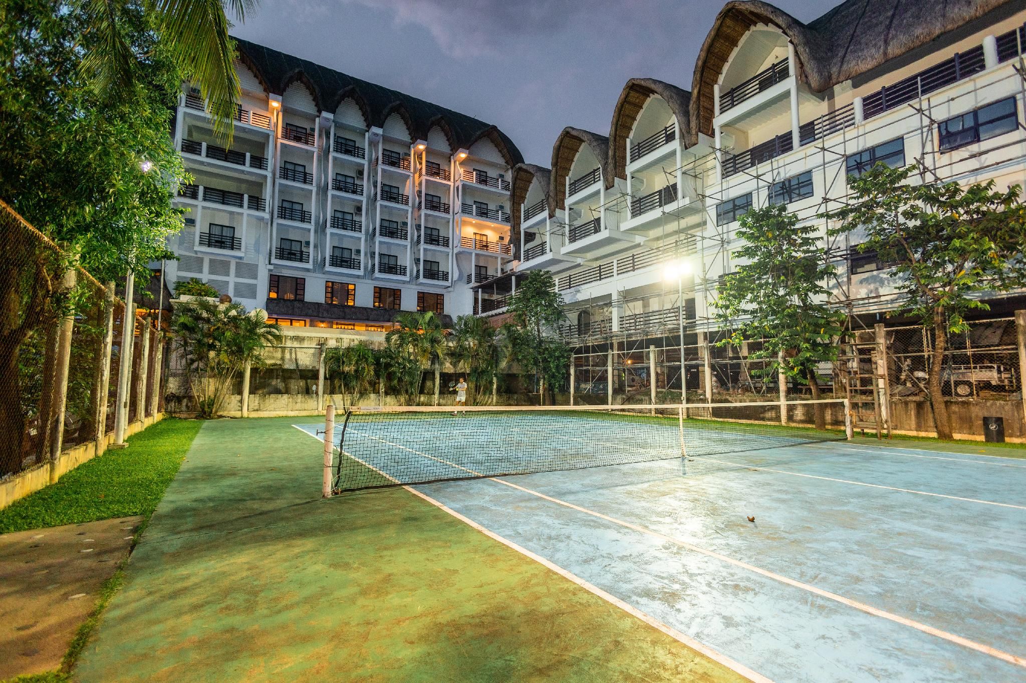 An outdoor tennis court with a net, surrounded by a building with multiple balconies and a fence.