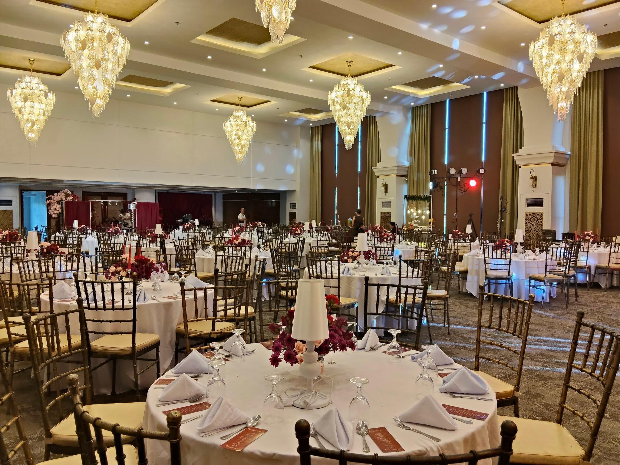 A banquet hall with round tables set for dining, large chandeliers, and a stage area.