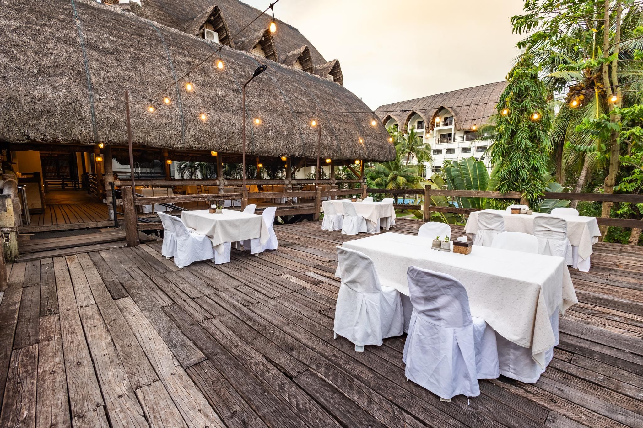 Un área de comedor al aire libre con estructuras de techo de paja, mesas y sillas de madera.