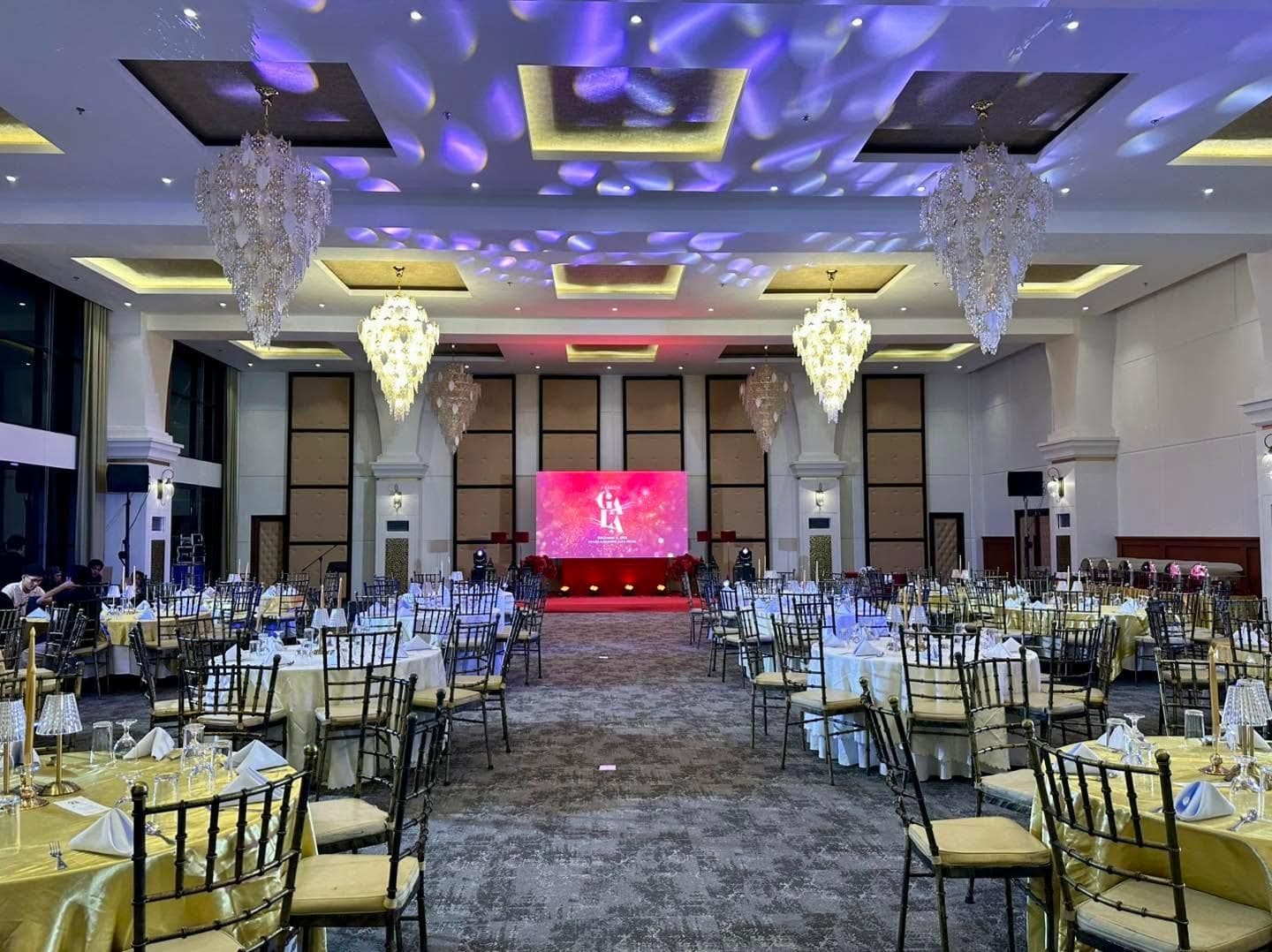A banquet hall with round tables, chairs, chandeliers, and a stage with a screen.