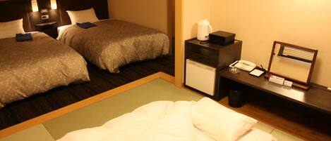 A hotel room with two beds, a desk with a telephone, a small refrigerator, and a bedside table with a lamp.