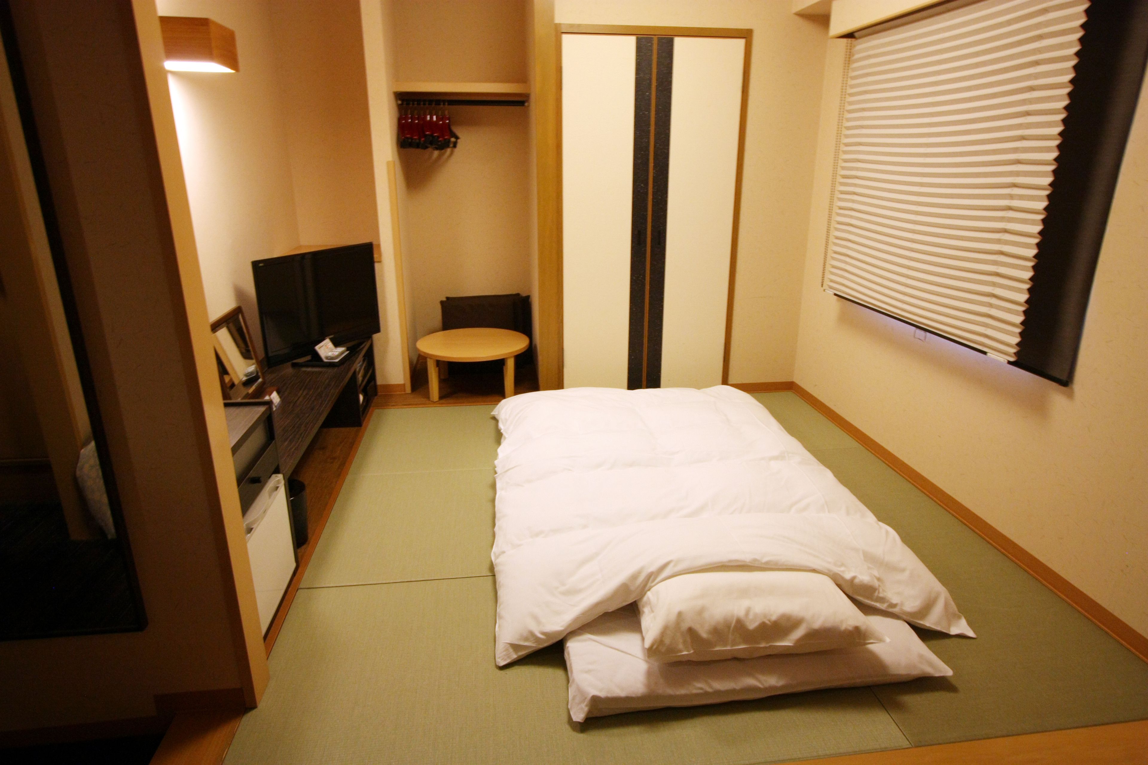 A room with tatami flooring, a bed with white bedding, a television on a stand, a small table, and a sliding door.