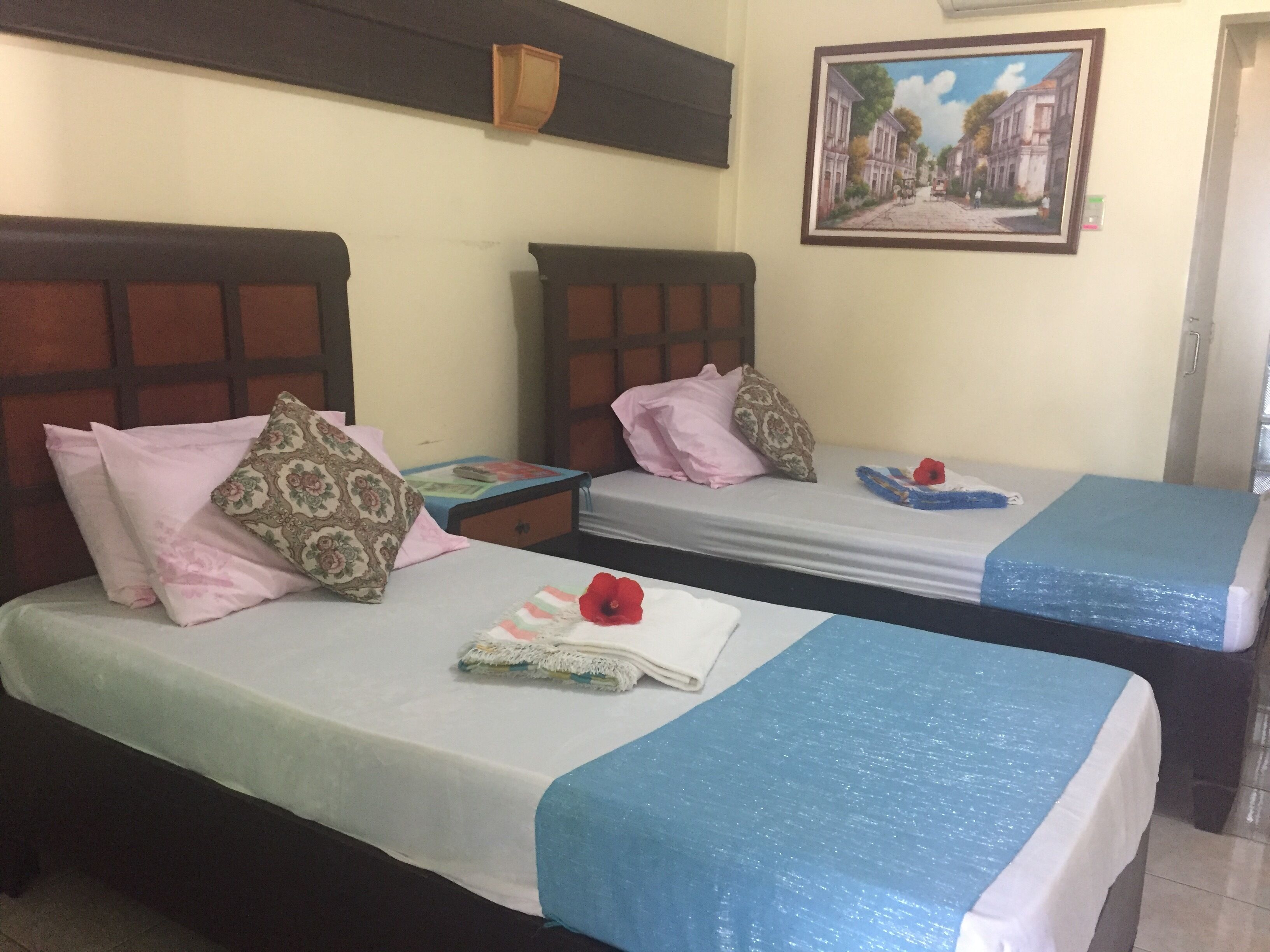 Habitación de hotel con dos camas individuales, cada una con una colcha azul y una almohada. Una mesita pequeña con un libro y una lámpara se encuentra entre las camas. Un cuadro cuelga de la pared sobre las camas.
