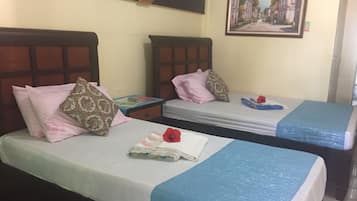 Habitación de hotel con dos camas individuales, cada una con una colcha azul y una almohada. Una mesita pequeña con un libro y una lámpara se encuentra entre las camas. Un cuadro cuelga de la pared sobre las camas.