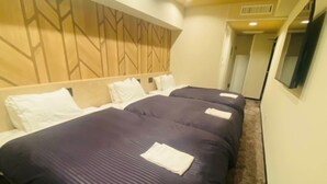 Habitació d'hotel amb tres llits, cadascun amb un llençol de llit blau fosc i almoades blanques. La habitació té un panell frontal de fusta amb un dibuix geomètric, un sòl amb moqueta i una televisió penjada a la paret.
