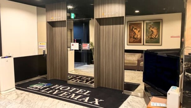 Un vestíbul d'hotel amb una taquilla d'adreça, una catifa negra i blanca amb el text 'HOTEL LIVEMAX', una televisió i obres d'art emmarcades a la paret.