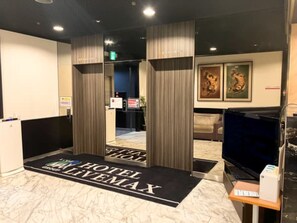 Um saguão de hotel com uma mesa de recepção, um tapete preto e branco com o texto 'HOTEL LIVEMAX', uma televisão e obras de arte emolduradas na parede.