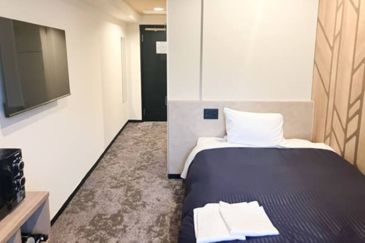 A hotel room with a bed, a flat-screen TV mounted on the wall, a wooden headboard, and a carpeted floor.