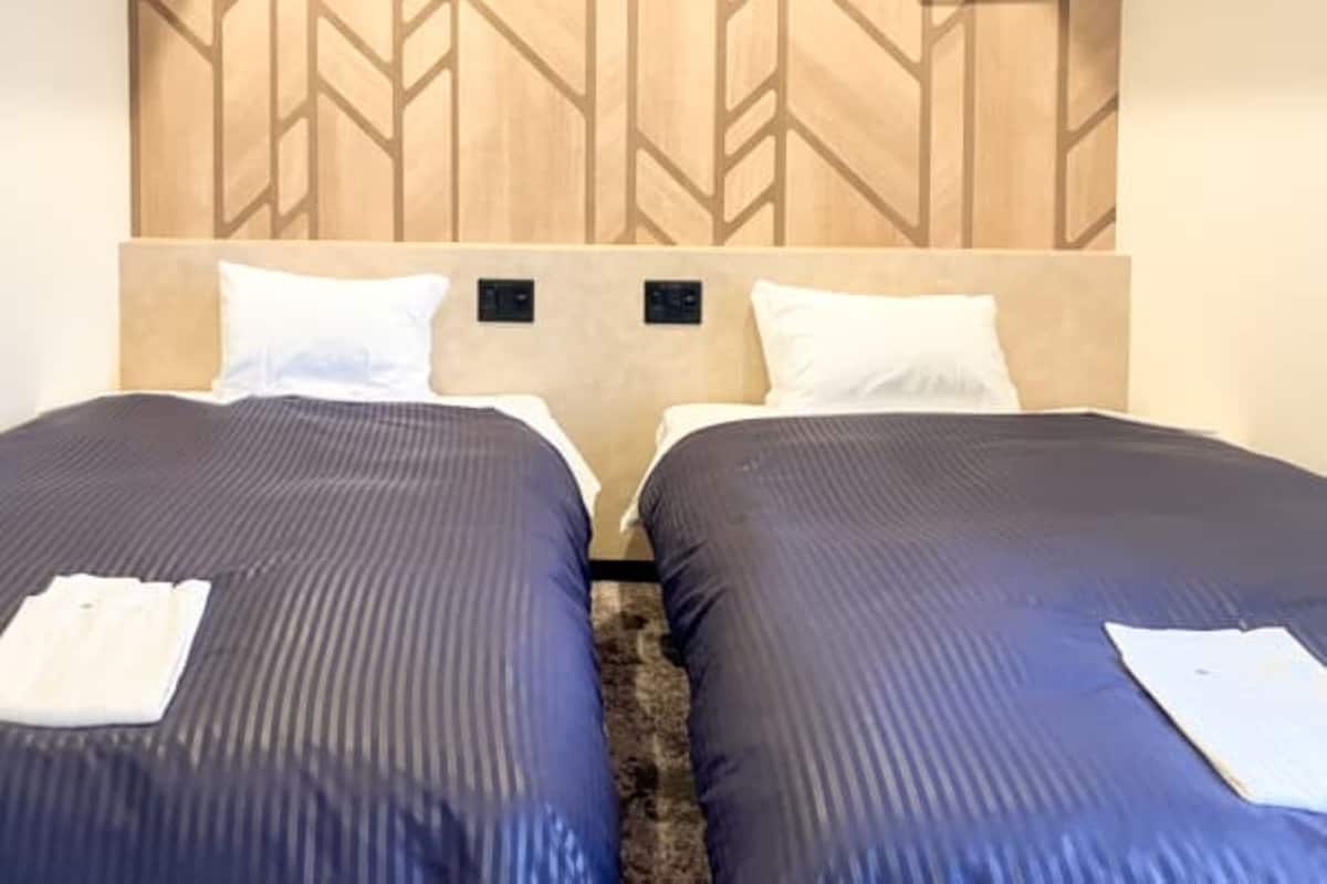 A hotel room with two beds, each with a dark blue bedspread and white pillows. The headboard has a geometric pattern, and there is a small shelf above the beds.