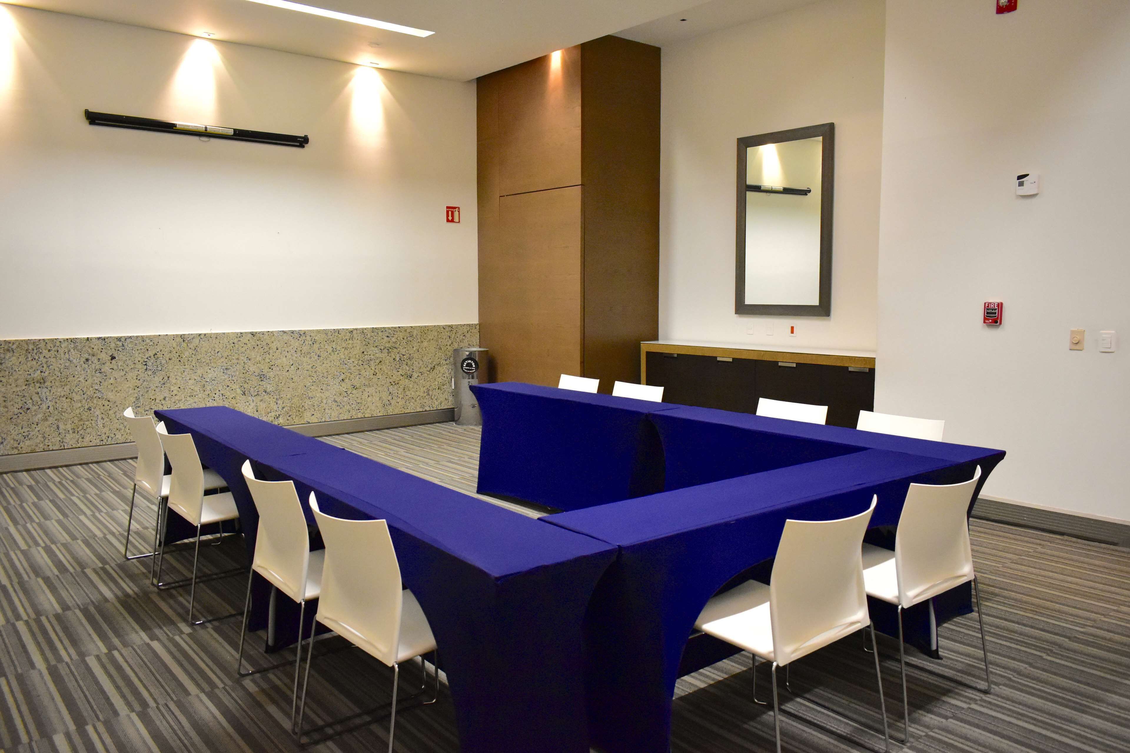 A conference room with a long table, white chairs, a blue tablecloth, a mirror, and a fire alarm on the wall.