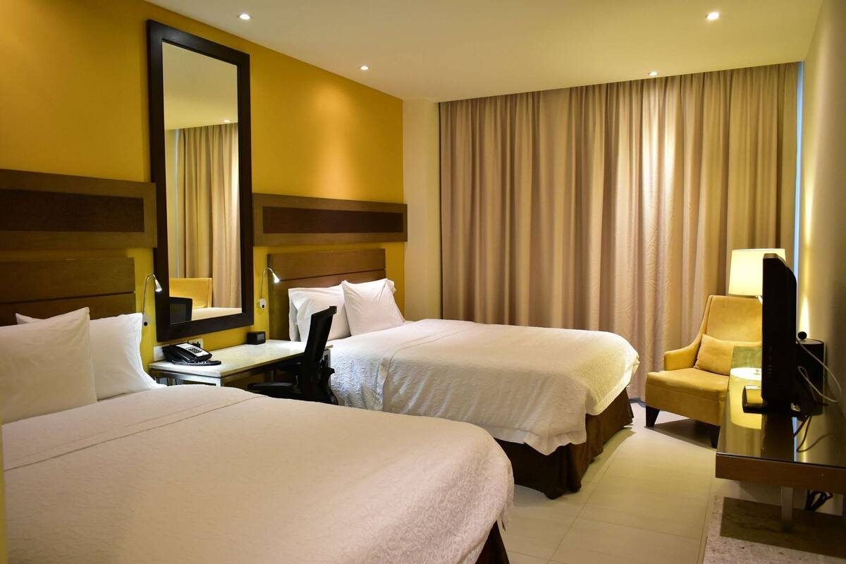 Habitación de hotel con dos camas, un escritorio con silla, televisión y un ventanal con cortinas.