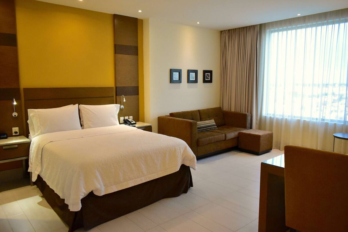 Una habitación de hotel con una cama grande, un sofá, una mesita y una ventana con cortinas.