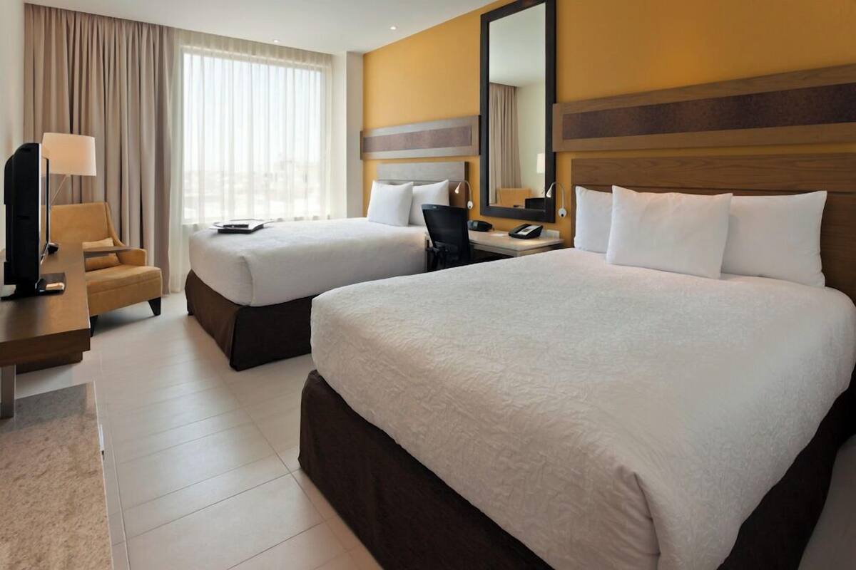 Habitación de hotel con dos camas, un escritorio, una silla, un televisor y un amplio ventanal con cortinas.