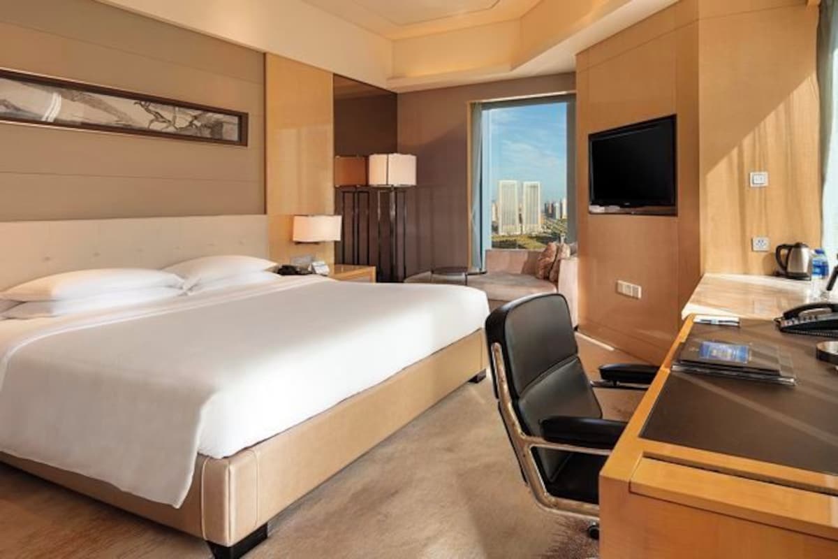 A hotel room with a large bed, a desk, a chair, a television, and a view of buildings.