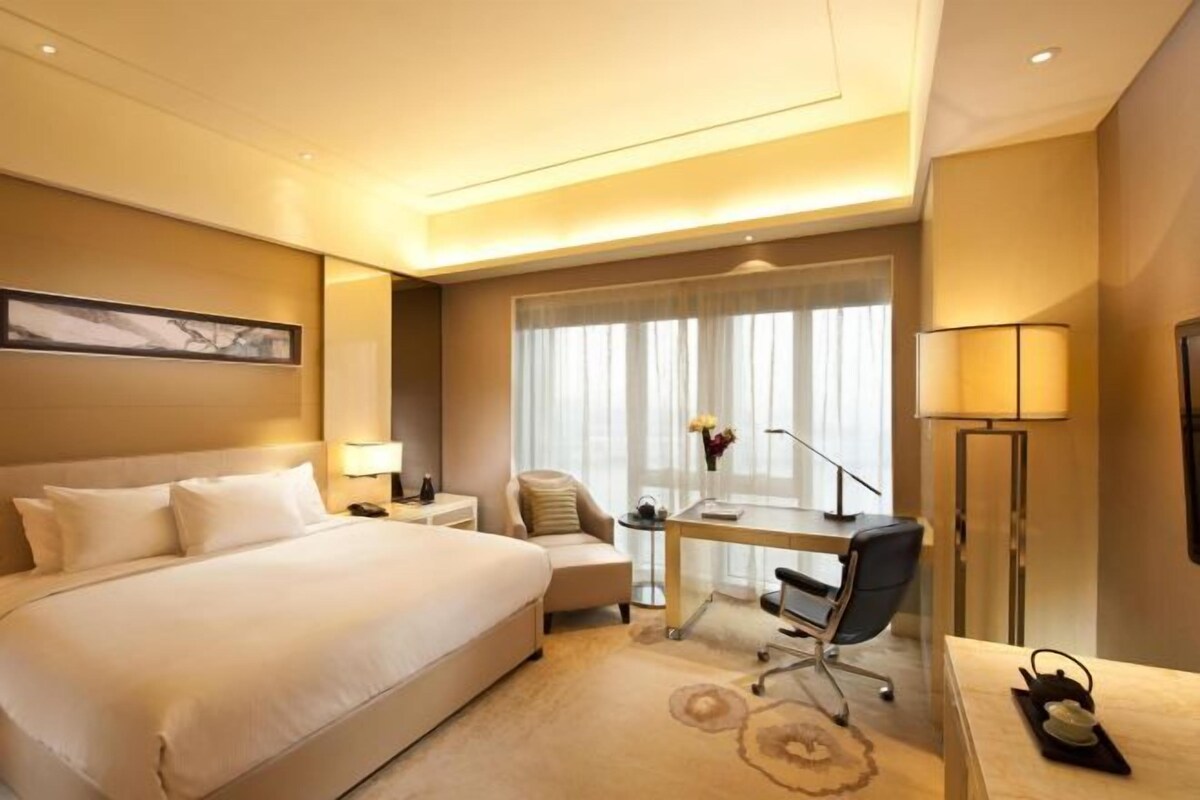 Habitación de hotel con una cama grande, un escritorio, una silla, una lámpara y vistas al exterior por la ventana.