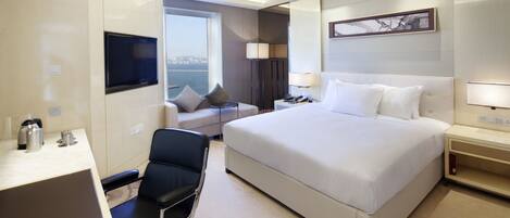 A hotel room with a large bed, a desk with a chair, a TV mounted on the wall, and a window offering a view of the city.