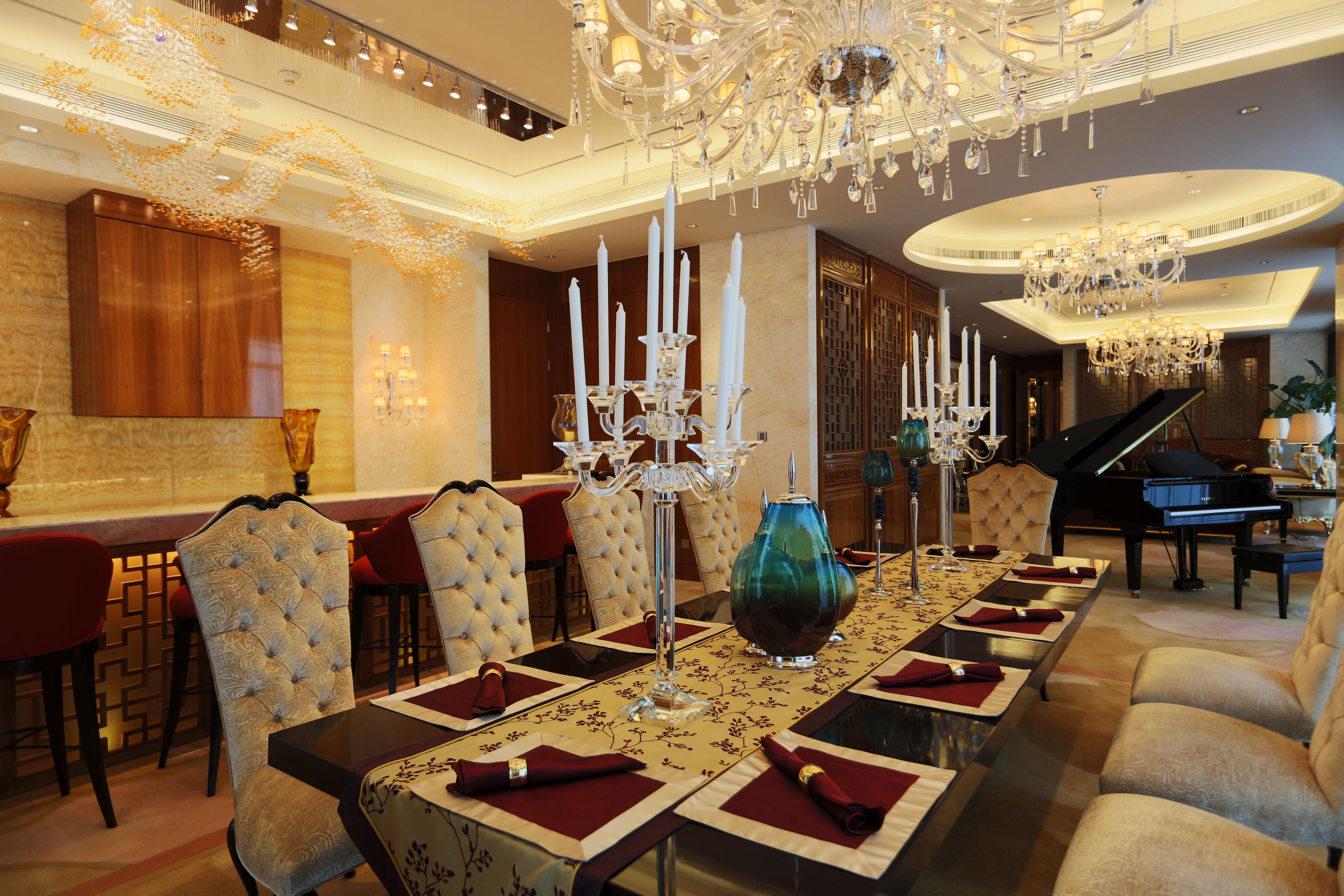 A luxurious dining room with a long table set for a meal, surrounded by plush chairs. A grand piano is visible in the background, indicating a possible entertainment area.