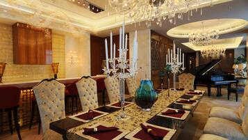 A luxurious dining room with a long table set for a meal, surrounded by plush chairs. A grand piano is visible in the background, indicating a possible entertainment area.