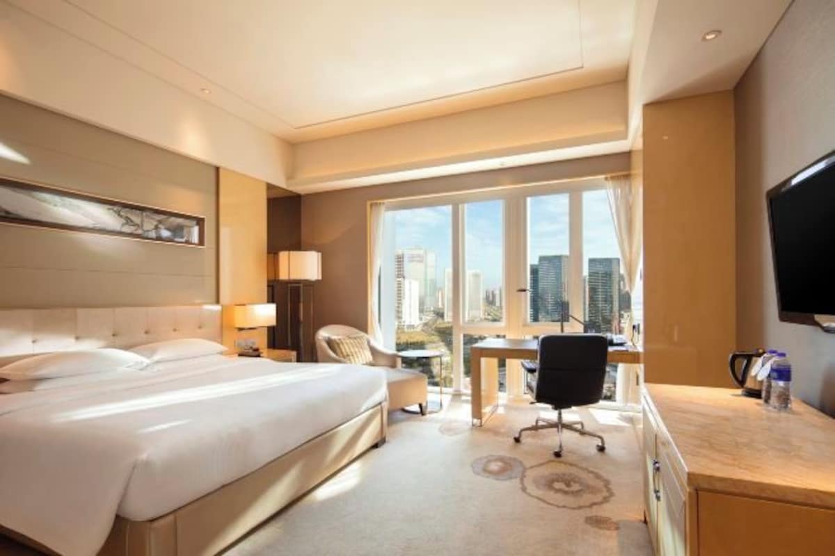 A hotel room with a large bed, a desk, a chair, a TV, and a view of the city.