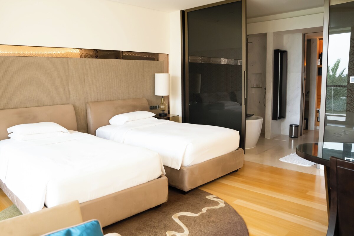 Une chambre d’hôtel avec deux lits, une table de chevet avec une lampe, une salle de bain avec des toilettes et un lavabo, et un balcon avec vue.