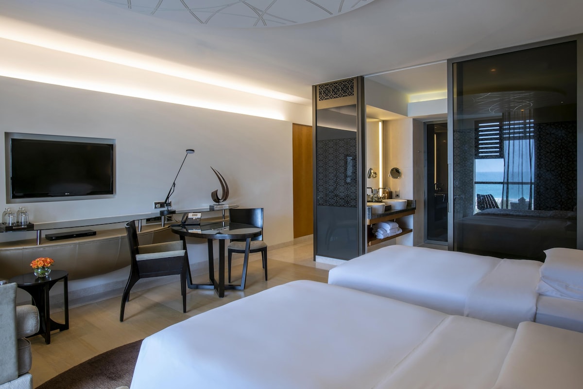 A modern hotel room with a large bed, a desk with a computer, a TV, and a bathroom with a large window.