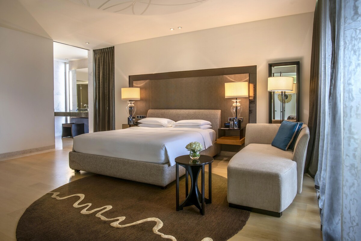 Une chambre d’hôtel avec un grand lit, deux tables de chevet avec des lampes, une petite table d’appoint, un canapé et un grand miroir.