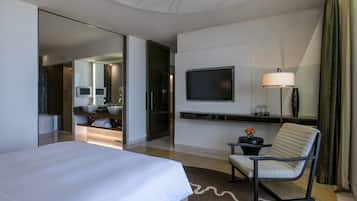 Quarto de hotel moderno com uma cama grande, televisão fixada na parede, área de estar com uma poltrona e uma pequena mesa com um vaso de flores.