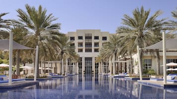 A swimming pool with lounge chairs and palm trees, surrounded by a building with multiple floors and balconies.