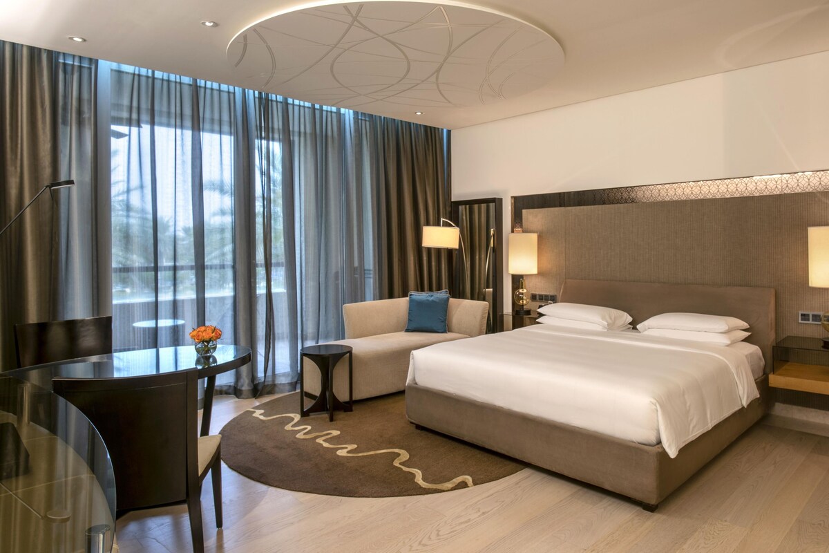 Une chambre d’hôtel moderne, dotée d’un grand lit, d’un canapé, d’un bureau avec une lampe et offrant une vue sur l’extérieur grâce à de grandes fenêtres.