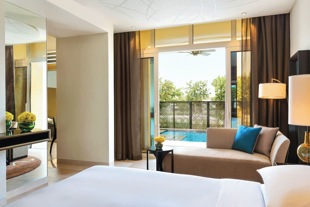 Une chambre d’hôtel avec un lit, un canapé, une petite table ornée d’un vase de fleurs, un bureau avec un miroir et une vue sur une piscine.
