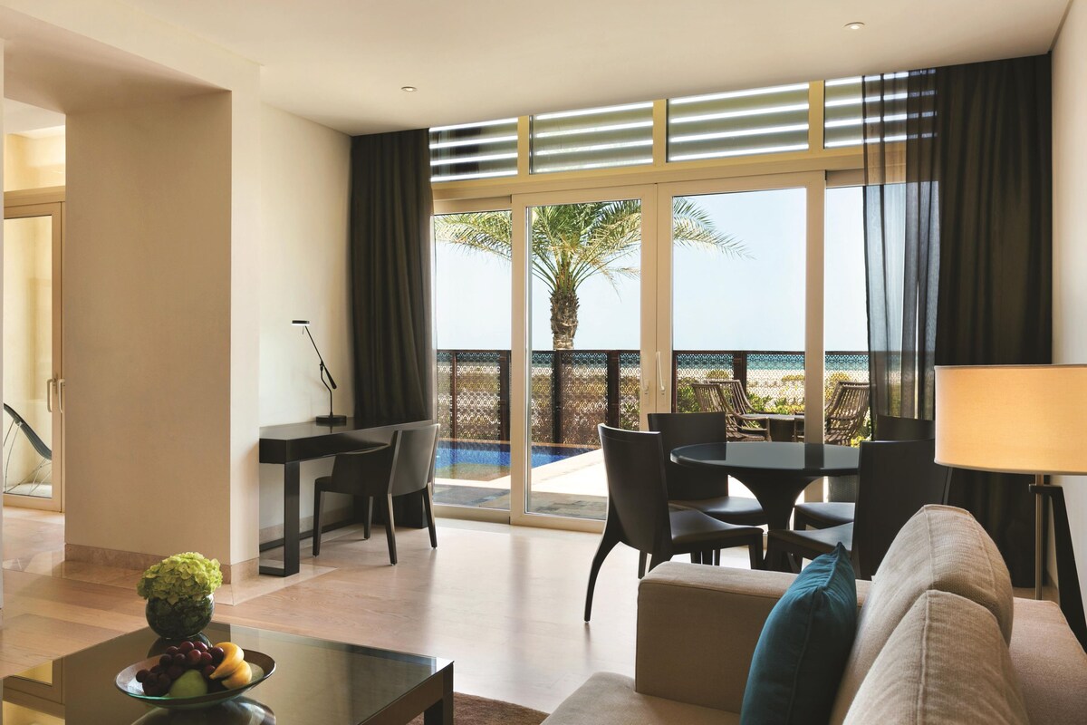 A modern hotel room with a dining area, a sofa, a table with fruit, and a view of the sea.