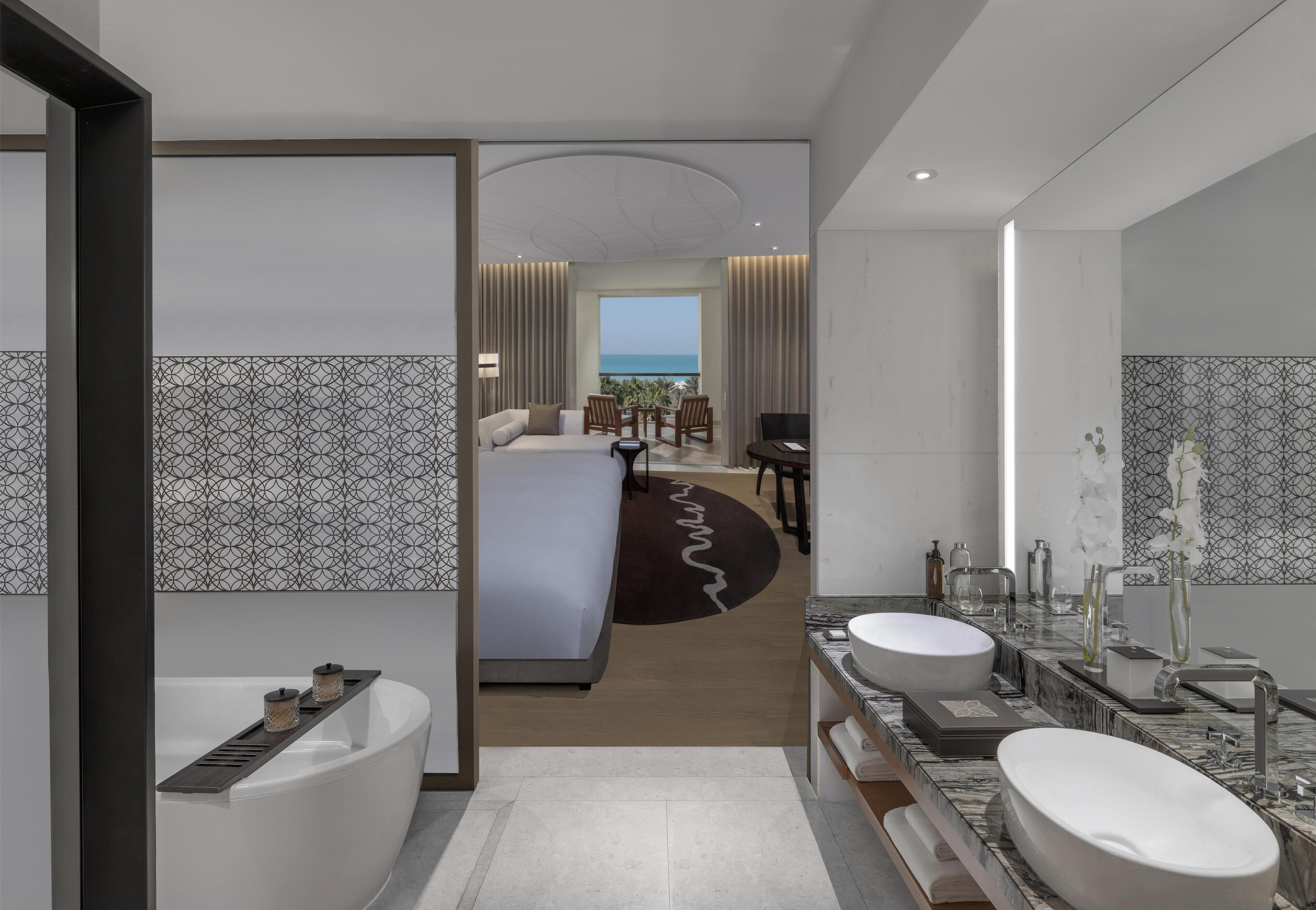 A modern hotel room with a large bed, a bathroom with dual sinks, and a view of the ocean.