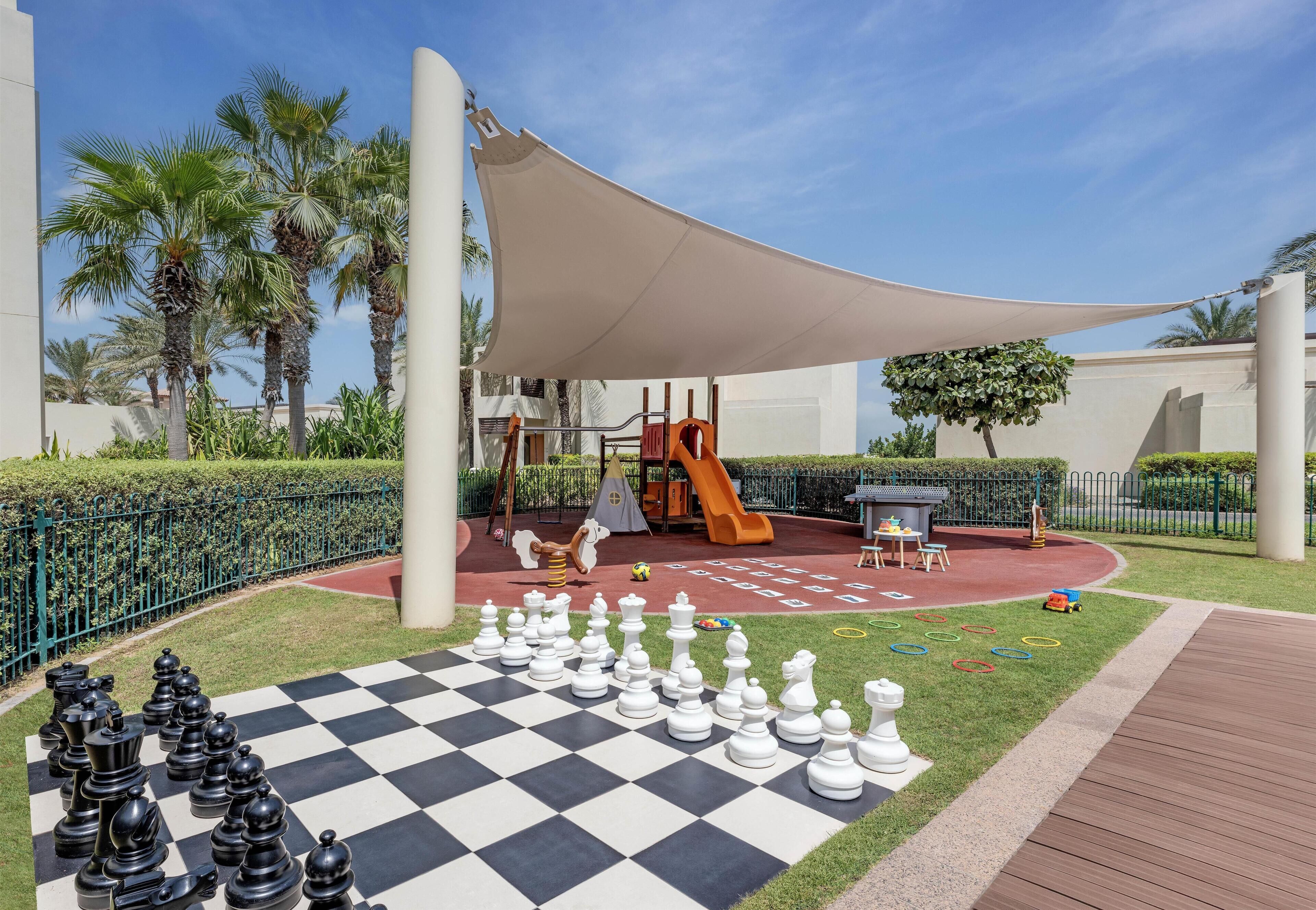 A checkered chessboard with chess pieces, a playground with a slide, palm trees, and a large canopy.