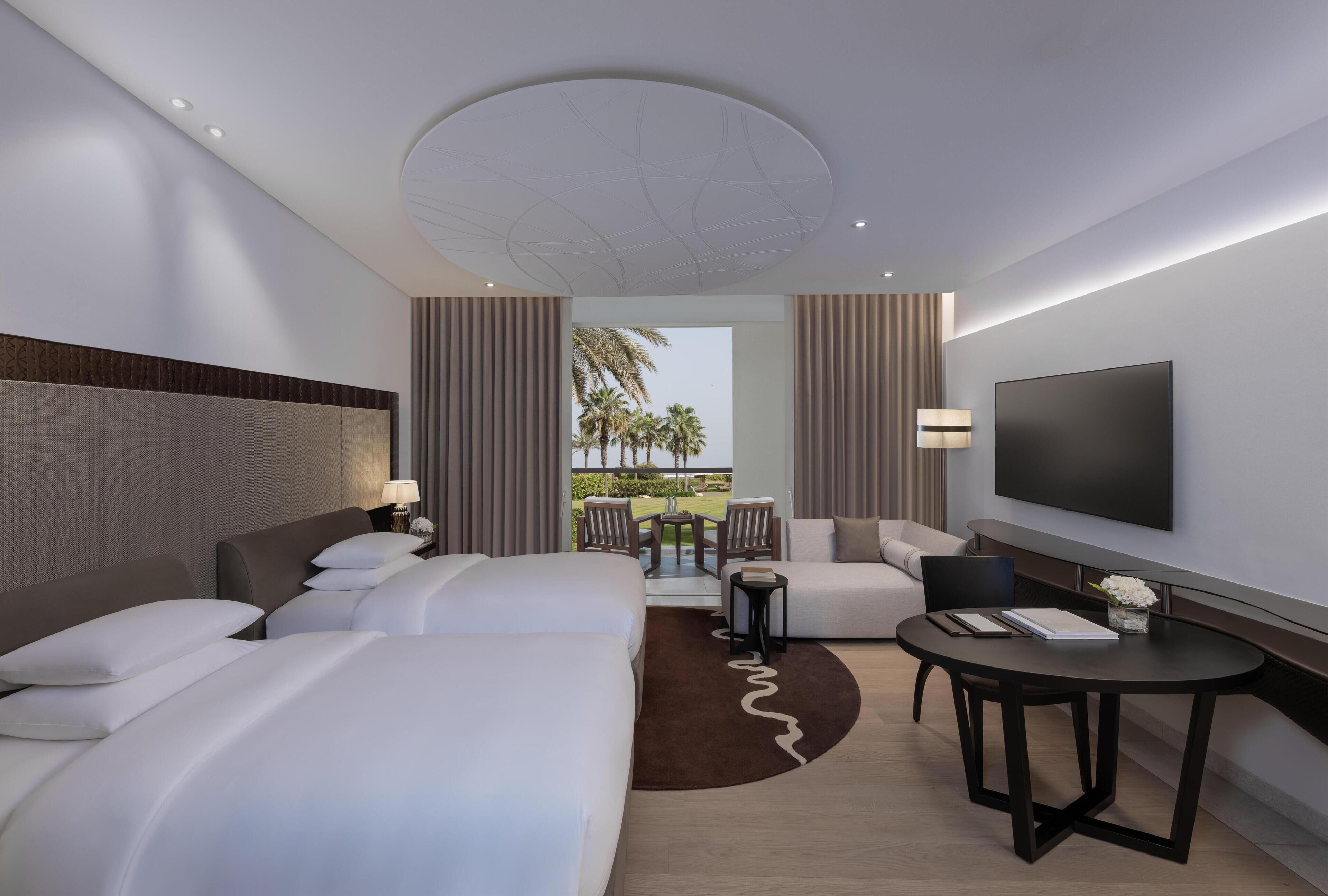 A hotel room with two beds, a TV, a sofa, a small table, and a balcony with a view of palm trees.