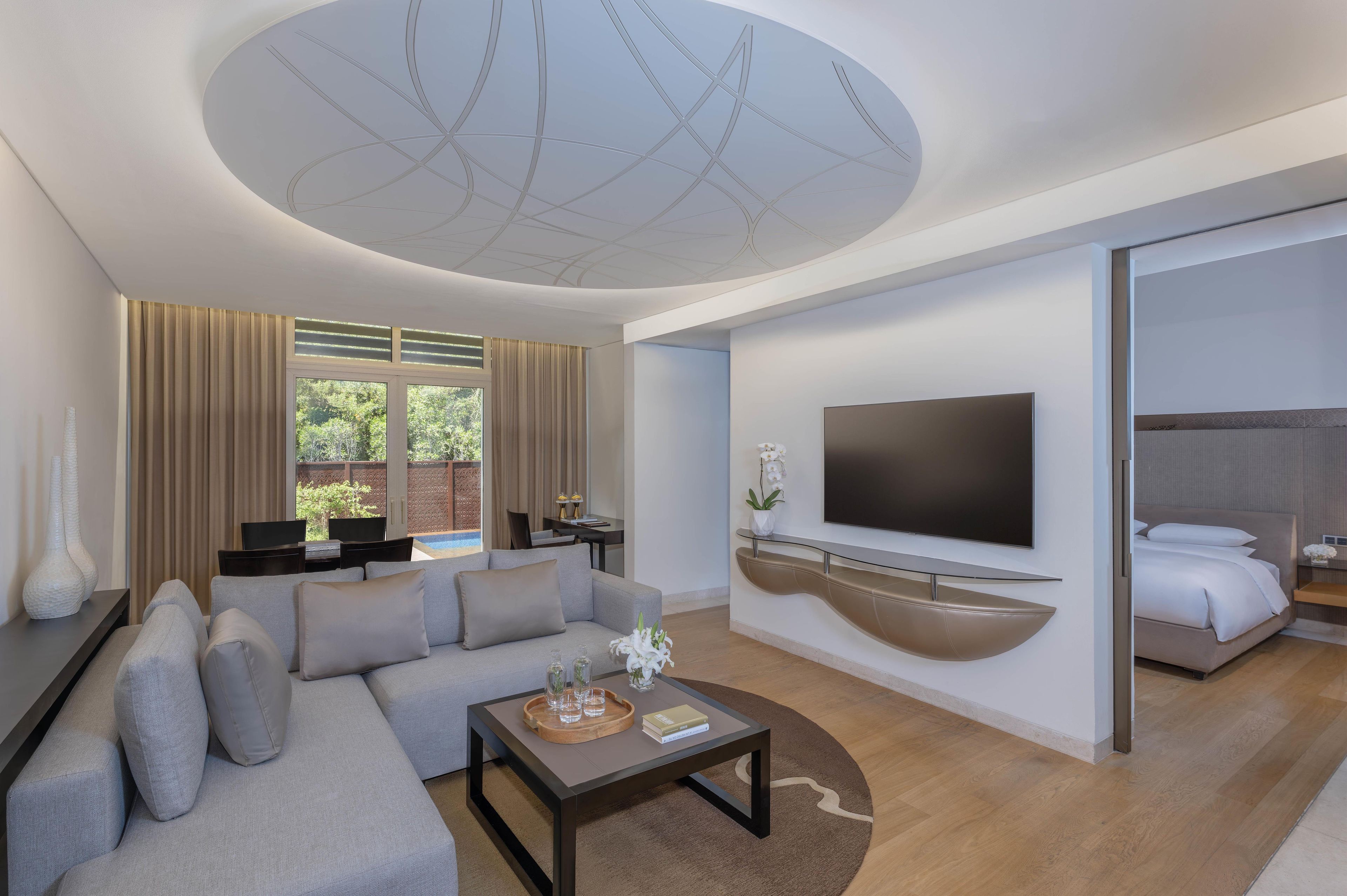 A modern hotel room with a sofa, a coffee table, a TV mounted on the wall, a bed, and a view of greenery outside.