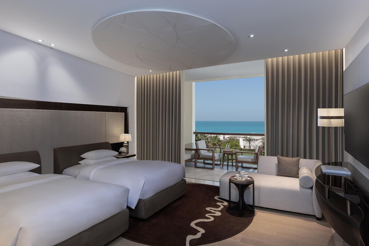 Une chambre d’hôtel avec deux lits, un canapé, une télévision et un balcon donnant sur la mer.