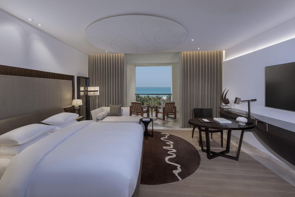 A hotel room with a large bed, a desk, a chair, a TV, and a view of the ocean.