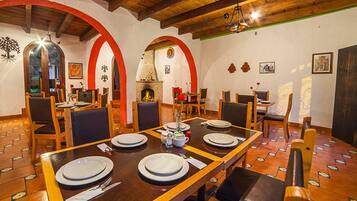A dining area with a long wooden table set for four, surrounded by chairs, and a fireplace in the background.