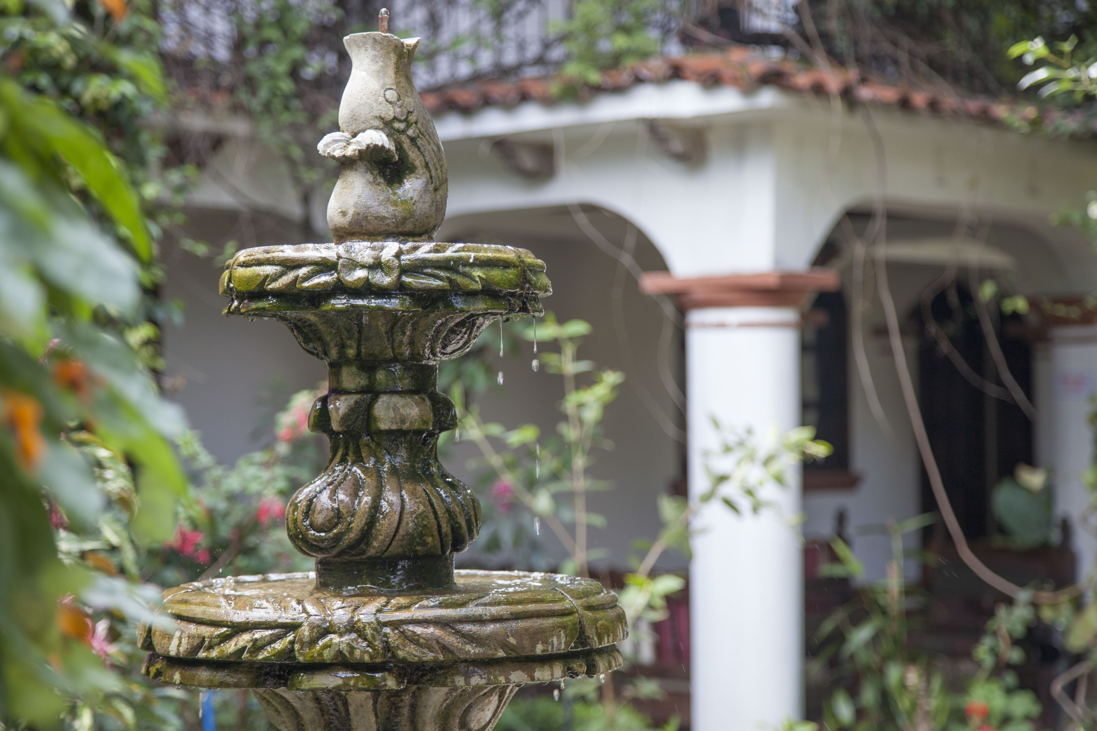 A stone fountain with a bird figure on top, surrounded by lush greenery and a white building with a tiled roof in the background.