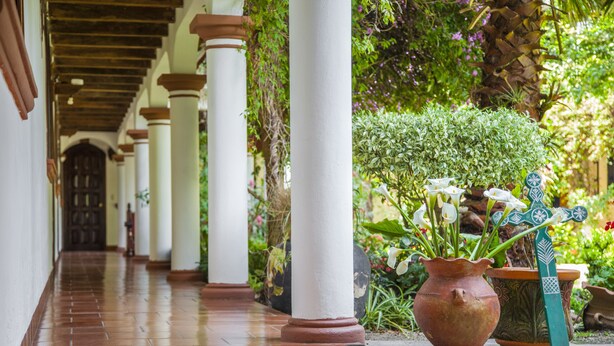 Un pasillo con columnas blancas, suelo de baldosas y plantas en macetas.