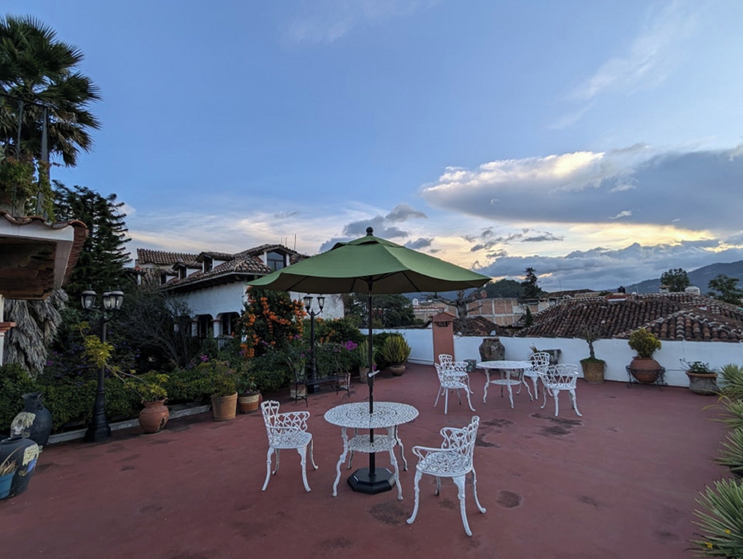 Un patio al aire libre con sillas y mesas de metal blanco, rodeado de plantas en macetas y un paraguas verde.