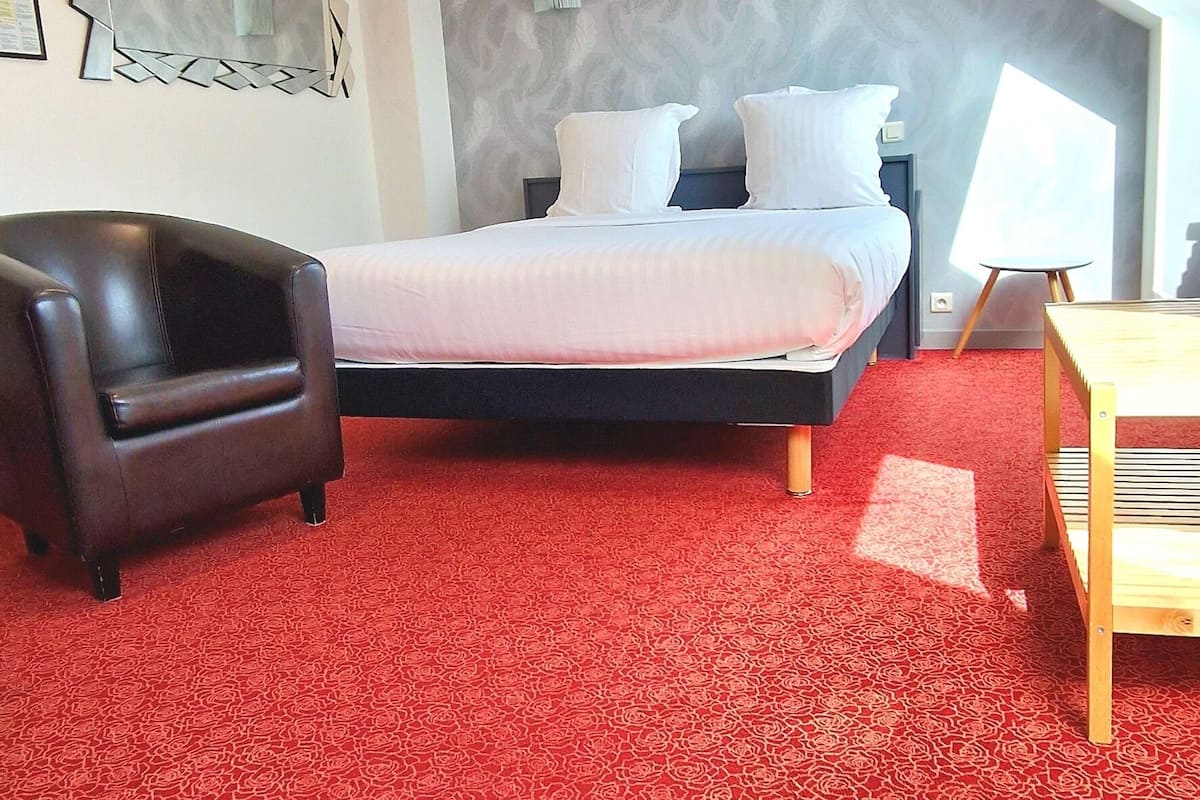 A bedroom with a red patterned carpet, a bed with white bedding, a dark leather chair, a wooden side table, and a large window providing natural light.