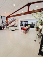 Amplio espacio interior con techo alto, escalera, mostrador de recepción y zona de comedor con mesa y sillas rojas.