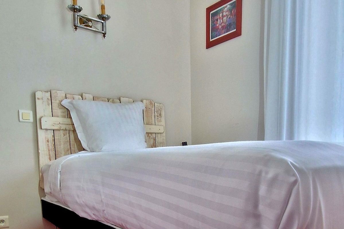 A hotel room with a bed, a red carpet, a white headboard, a wall-mounted light fixture, and a framed picture.