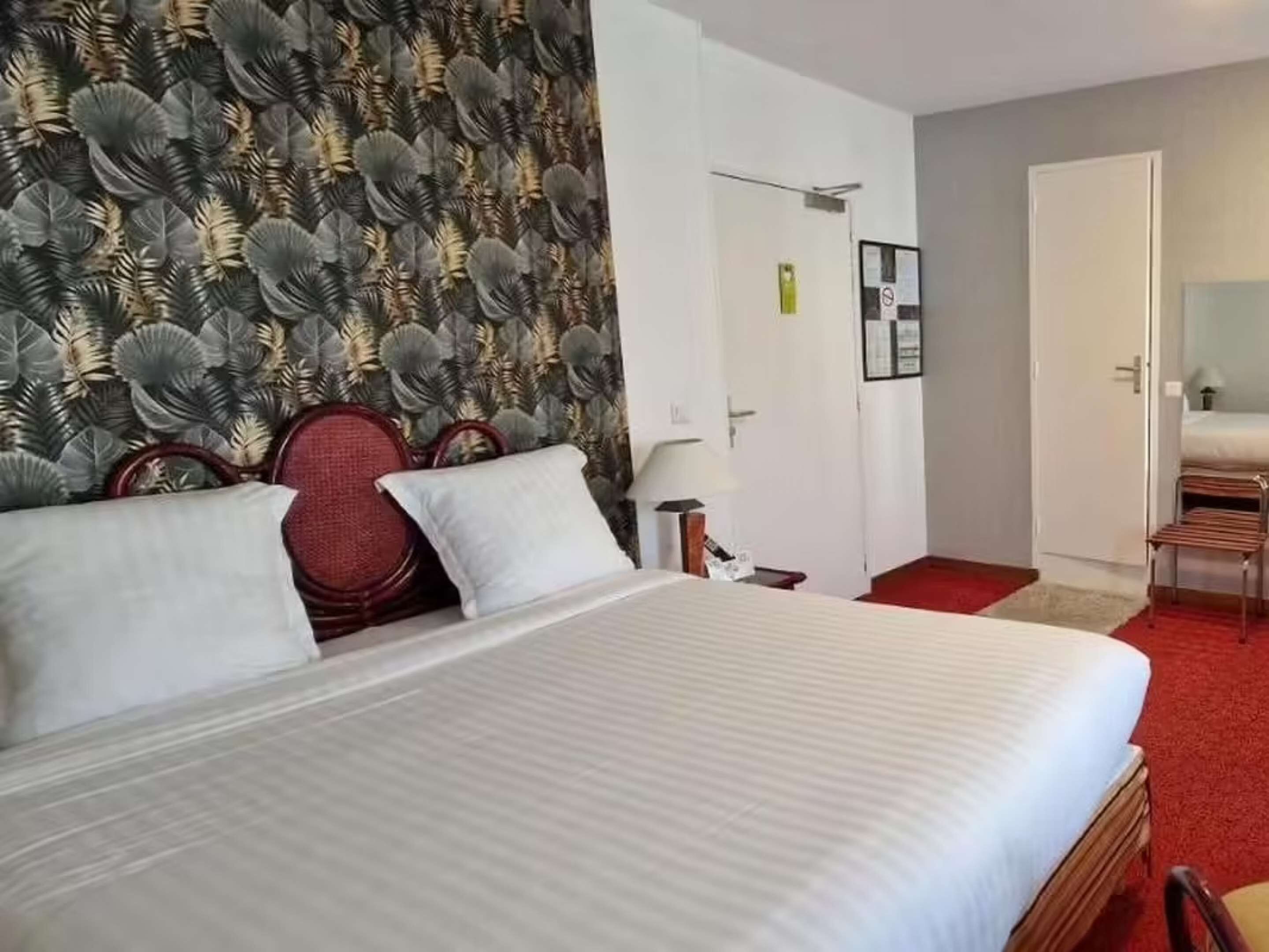 Una camera d'albergo con un letto, una sedia rossa, un comodino in legno e una porta.