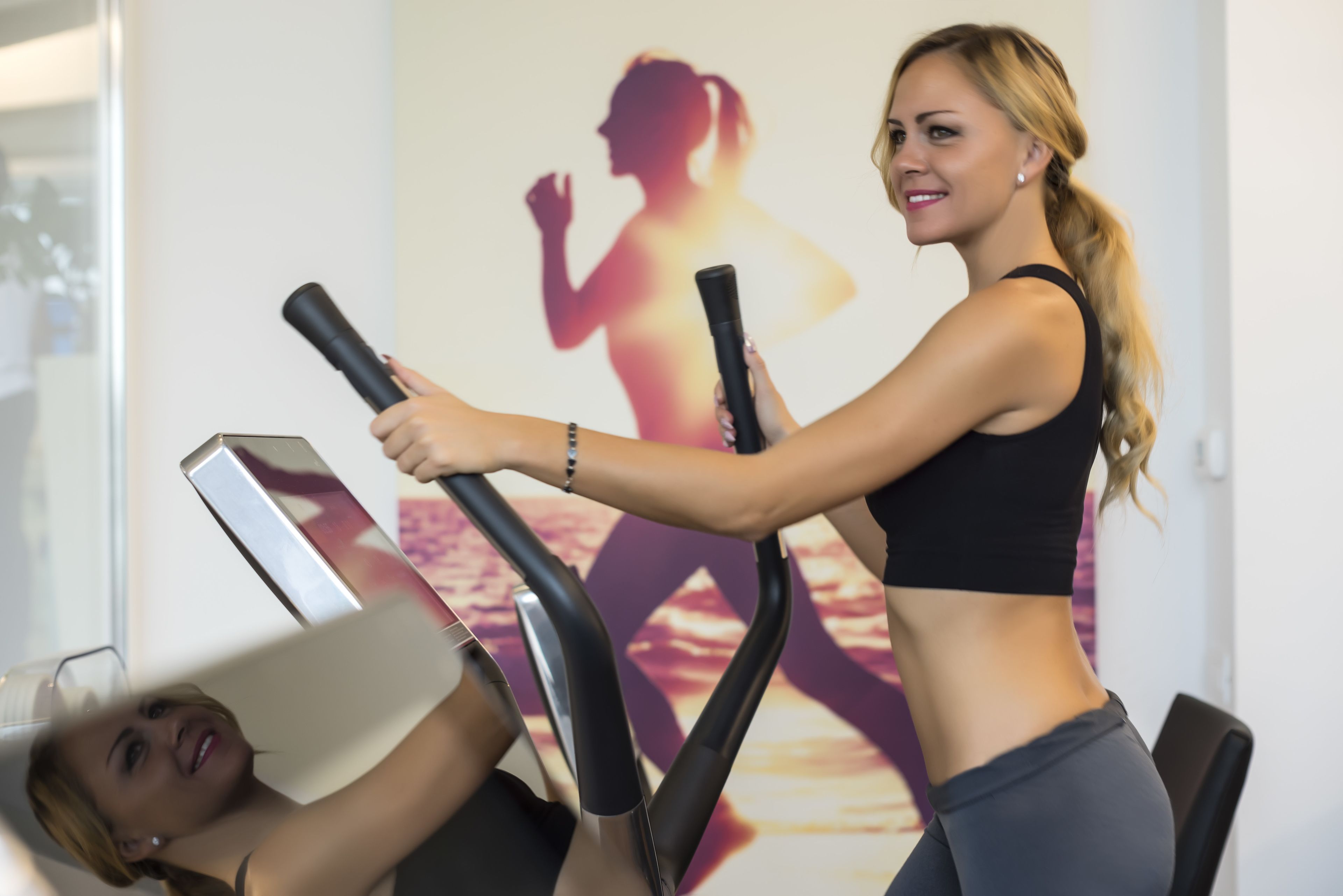 A woman using an elliptical machine in a gym with a poster of a woman exercising in the background.